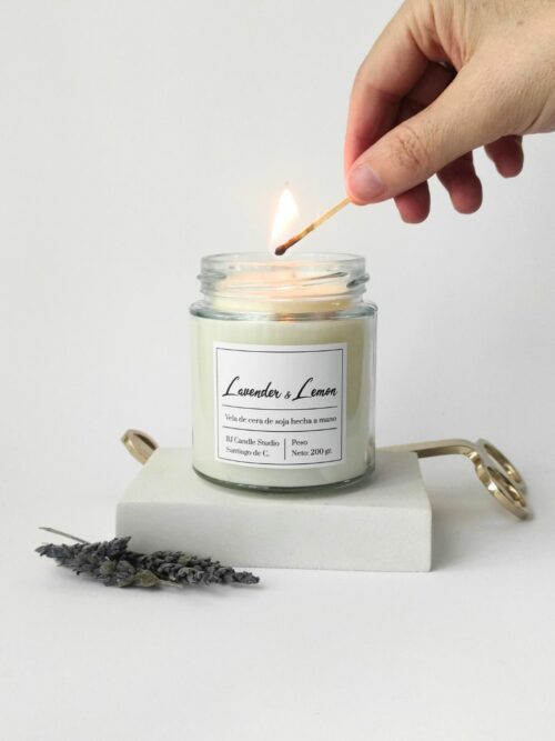vela siendo encendida por una mano. Al lado hay unas flores secas de lavanda como decoración y en la parte de atrás, un cortamechas dorado.