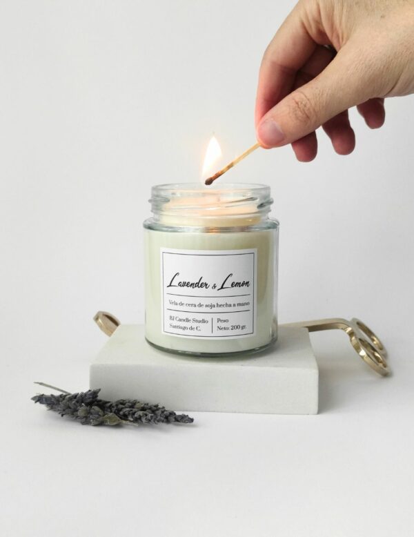 vela siendo encendida por una mano. Al lado hay unas flores secas de lavanda como decoración y en la parte de atrás, un cortamechas dorado.