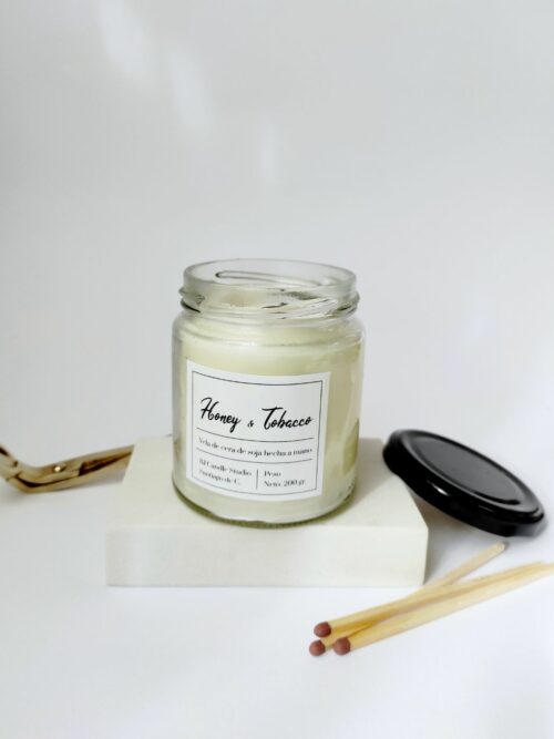 Vela aromática Miel y Tabaco hecha a mano con cera de soja. Tiene la tapa negra al lado y unas cerillas largas decorando. Estilo minimalista.