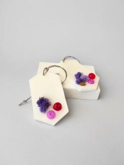 dos ambientadores sólidos colocados sobre un soporte. Están decorados con una flor seca de color violeta y dos bolitas de cera una roja y otra de color rojo. Aroma a Clean Cotton