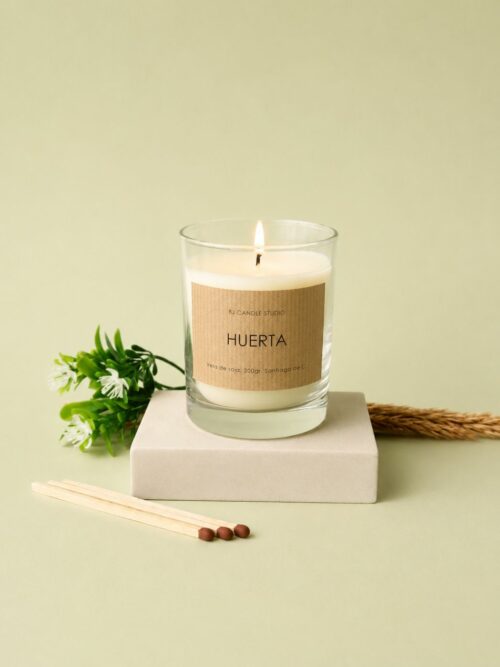 vela aromática con pegatina kraft. Escena minimalista con unas hojas verdes decorativas detrás y unas cerillas al lado.